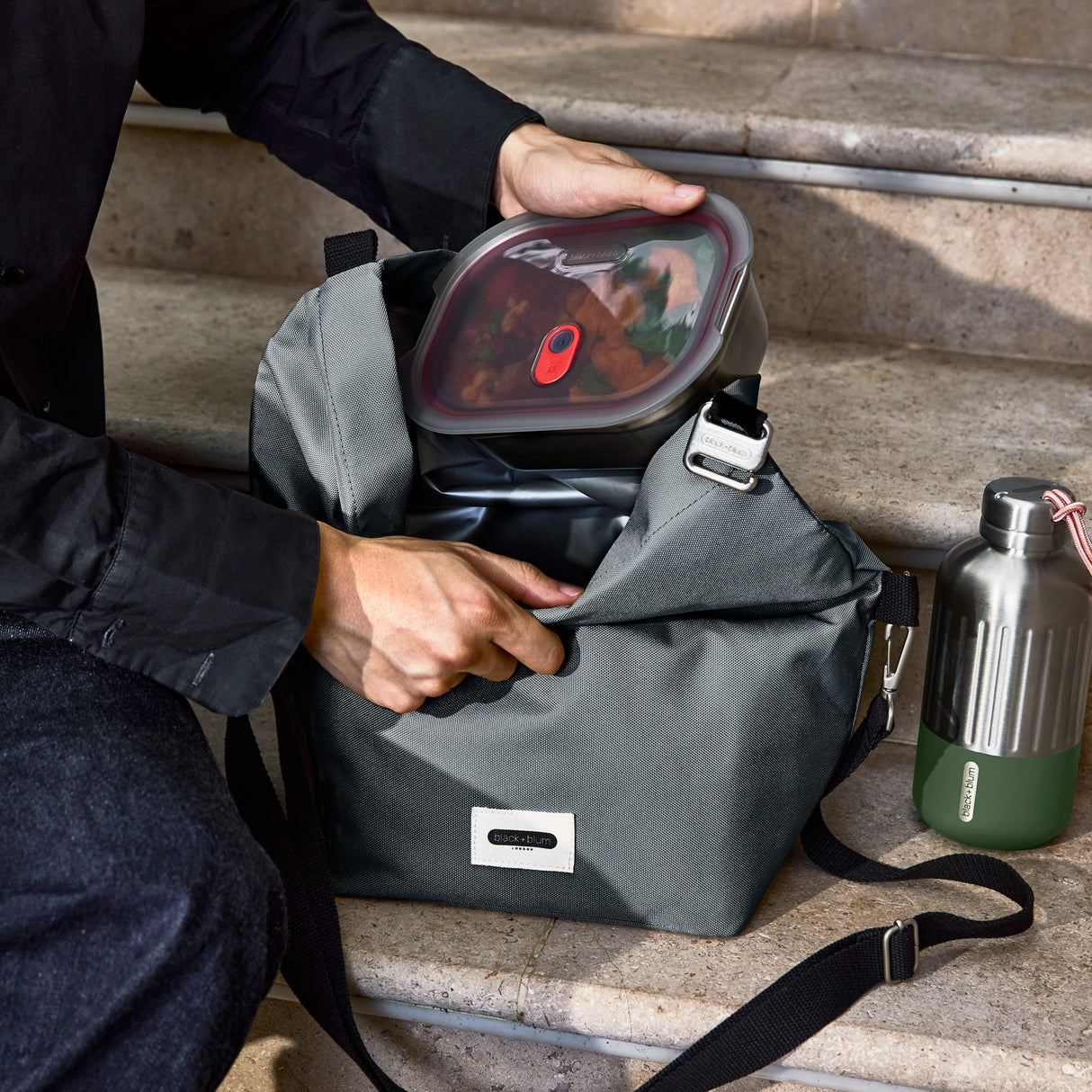 Black+Blum insulated lunchbag in slate color, large size with a sleek design, shown on stairs in a vertical view.
